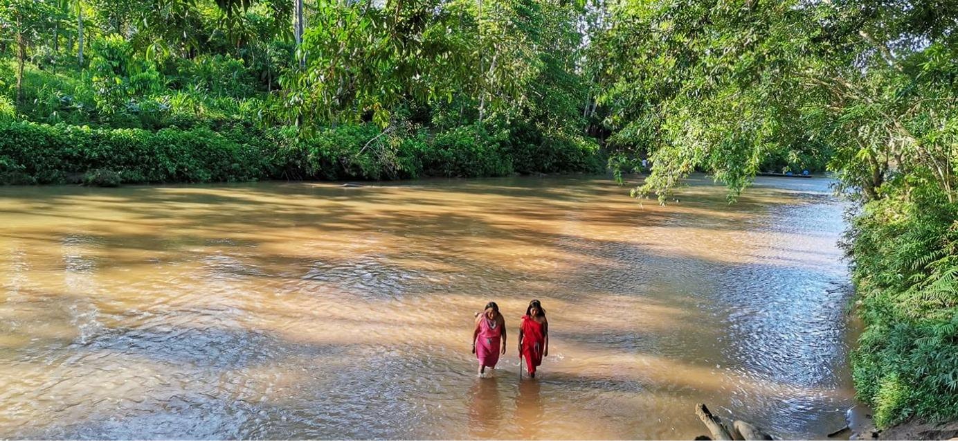 Dos mujeres con vestidos rojos cruzan a pie un río de la Amazonía, rodeado de bosque.